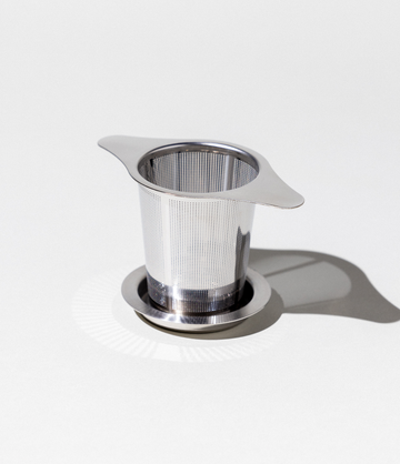 A stainless steel tea infuser with a handle and basket design, placed on a white surface.